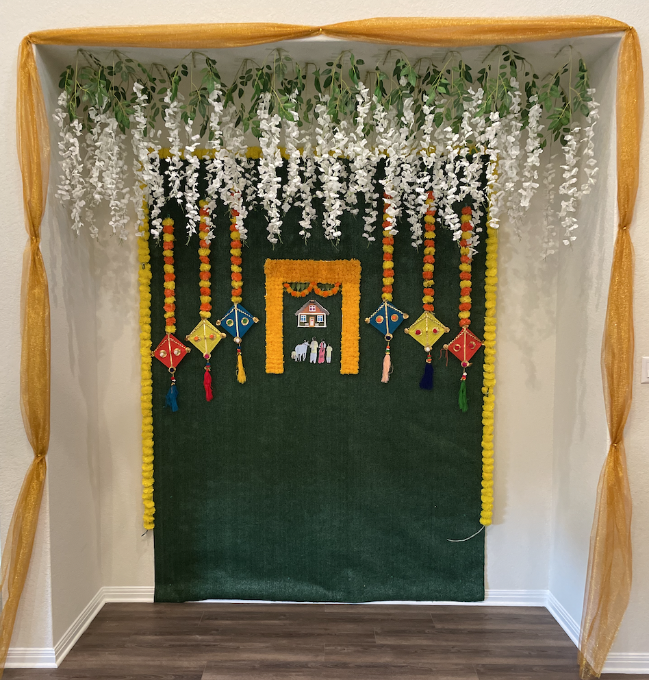 Decorative event backdrop featuring a green fabric with floral garlands, colorful kites, and an artistic depiction of a house.